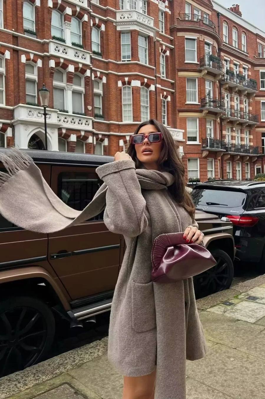 Khaki - A woman wearing a taupe brushed wool mid-length coat with an integrated matching scarf, styled with dark sunglasses and a plum clutch against a classic brick building background.