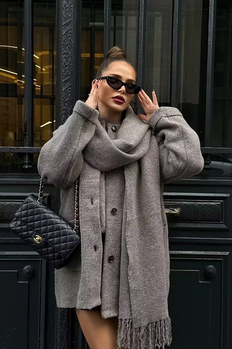 Grey - A woman wearing a taupe brushed wool mid-length coat with an integrated matching scarf, styled with dark sunglasses and a plum clutch against a classic brick building background.