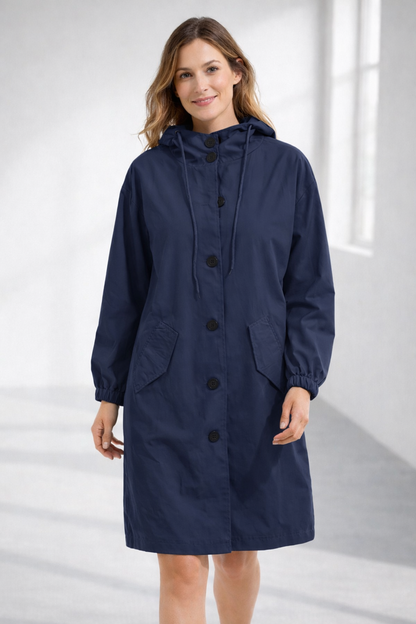 Navy blue - A woman wearing a teal mid-length hooded coat with a contrast black button-down front, angled flap pockets, and elasticated cuffs, set against a bright minimalist interior.