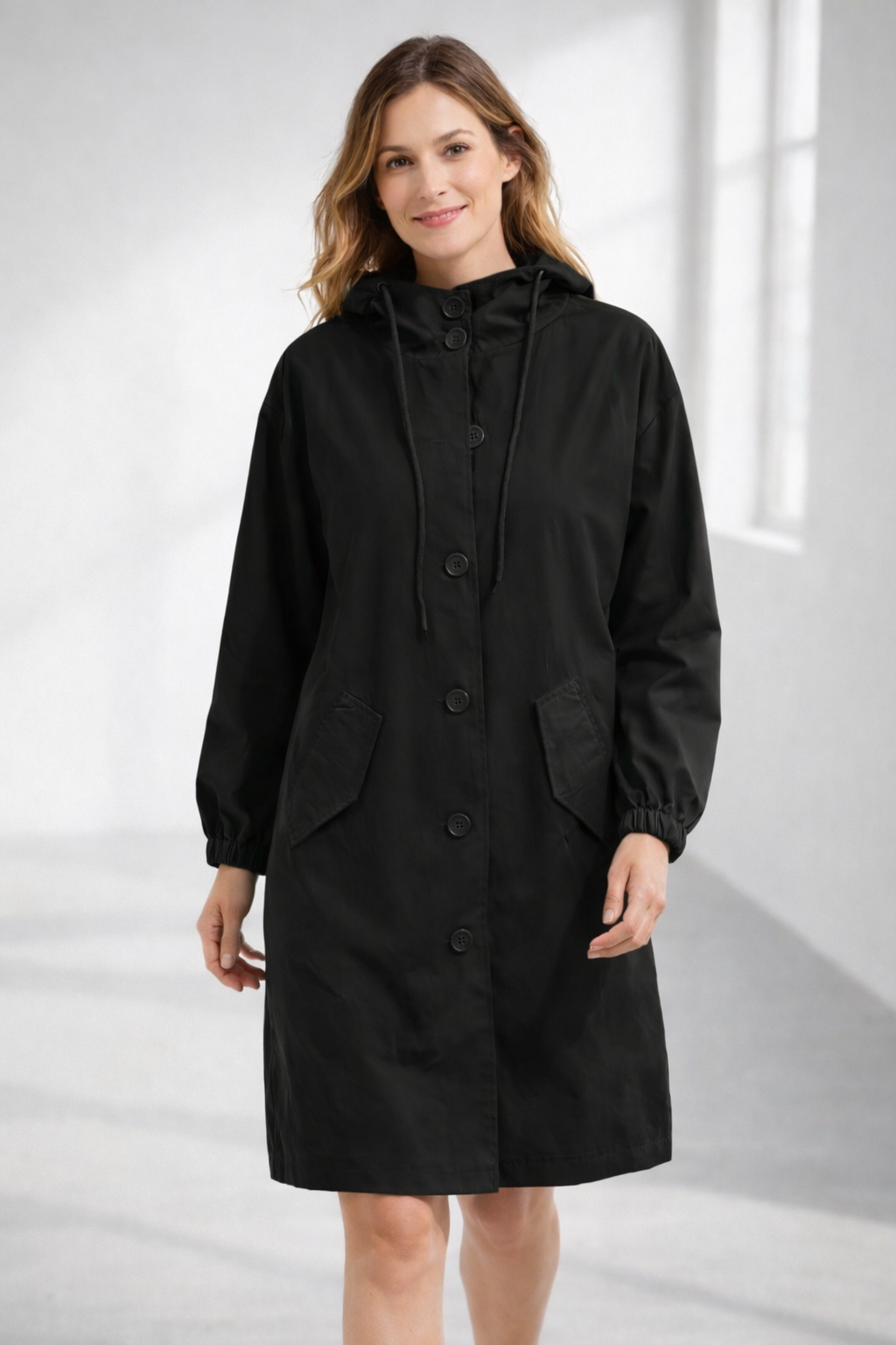 Black - A woman wearing a teal mid-length hooded coat with a contrast black button-down front, angled flap pockets, and elasticated cuffs, set against a bright minimalist interior.