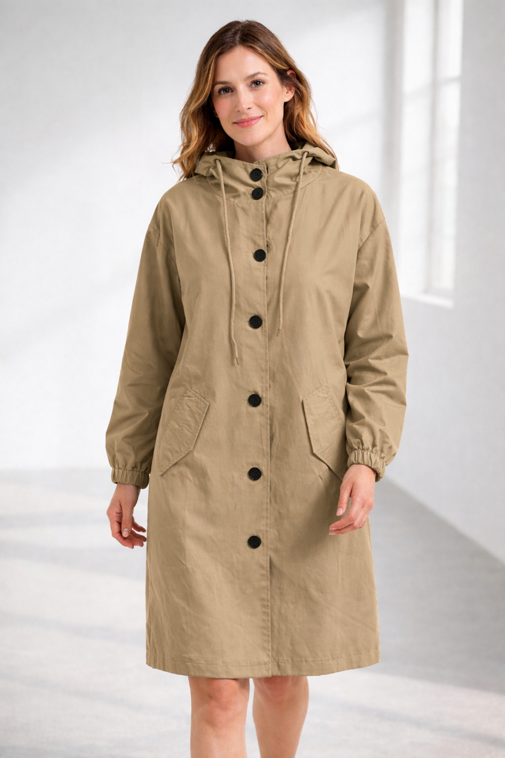 Beige - A woman wearing a teal mid-length hooded coat with a contrast black button-down front, angled flap pockets, and elasticated cuffs, set against a bright minimalist interior.
