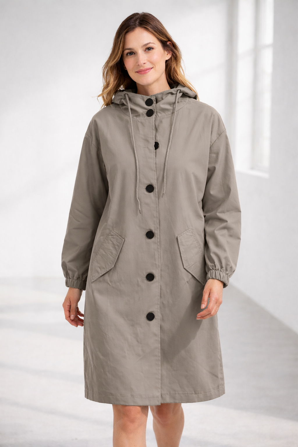 Apricot - A woman wearing a teal mid-length hooded coat with a contrast black button-down front, angled flap pockets, and elasticated cuffs, set against a bright minimalist interior.