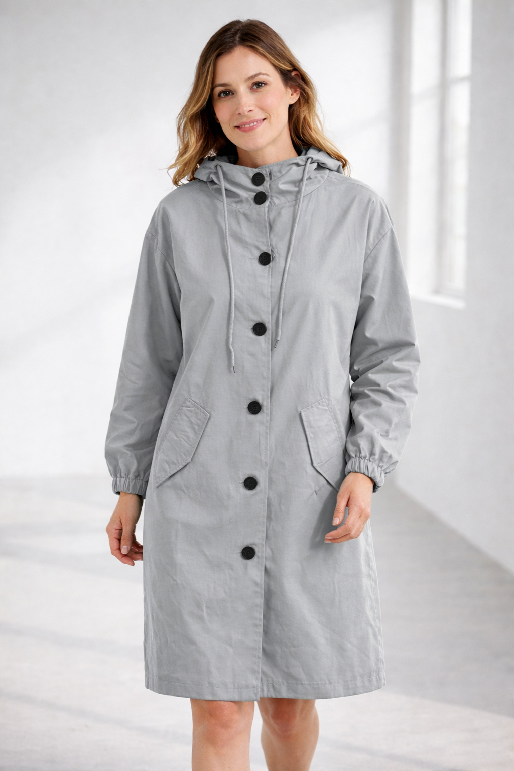 Grey - A woman wearing a teal mid-length hooded coat with a contrast black button-down front, angled flap pockets, and elasticated cuffs, set against a bright minimalist interior.