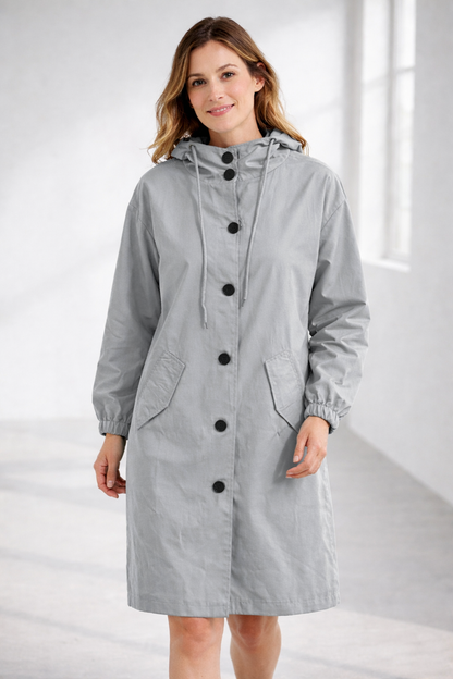 Grey - A woman wearing a teal mid-length hooded coat with a contrast black button-down front, angled flap pockets, and elasticated cuffs, set against a bright minimalist interior.