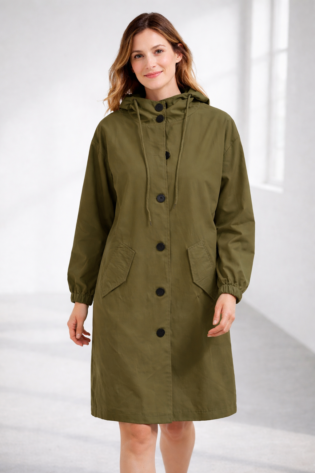Army green - A woman wearing a teal mid-length hooded coat with a contrast black button-down front, angled flap pockets, and elasticated cuffs, set against a bright minimalist interior.
