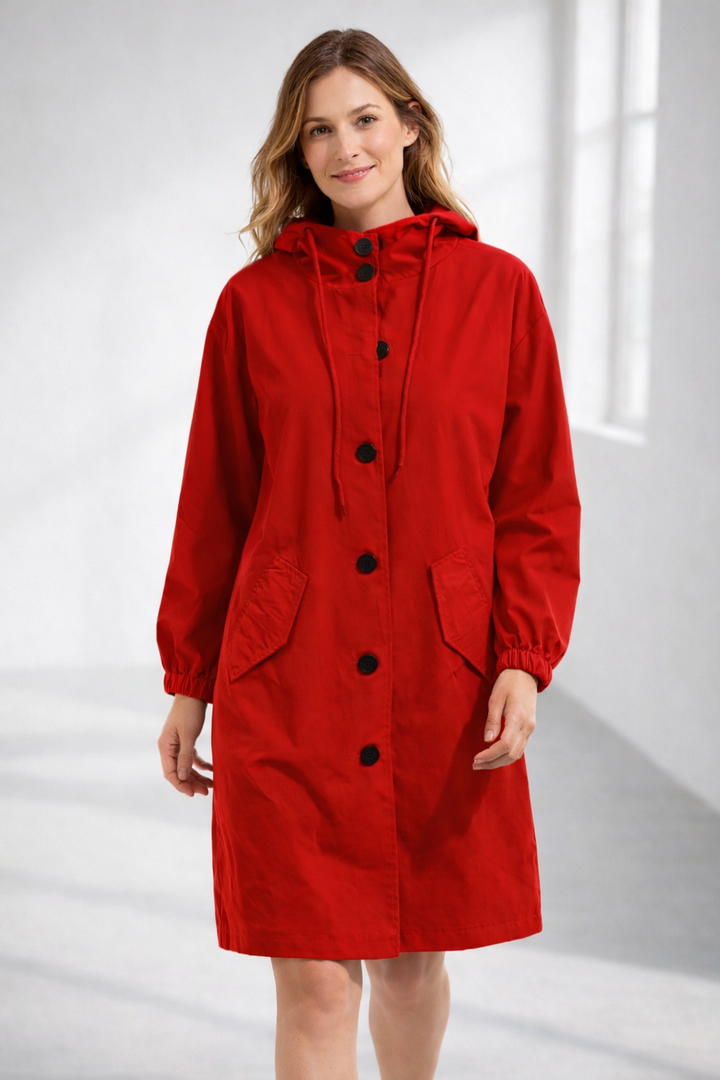Red - A woman wearing a teal mid-length hooded coat with a contrast black button-down front, angled flap pockets, and elasticated cuffs, set against a bright minimalist interior.