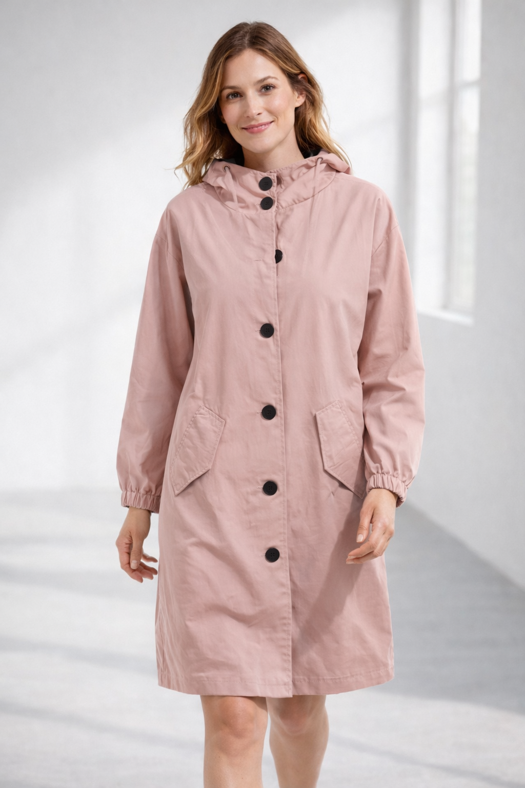 Pink - A woman wearing a teal mid-length hooded coat with a contrast black button-down front, angled flap pockets, and elasticated cuffs, set against a bright minimalist interior.