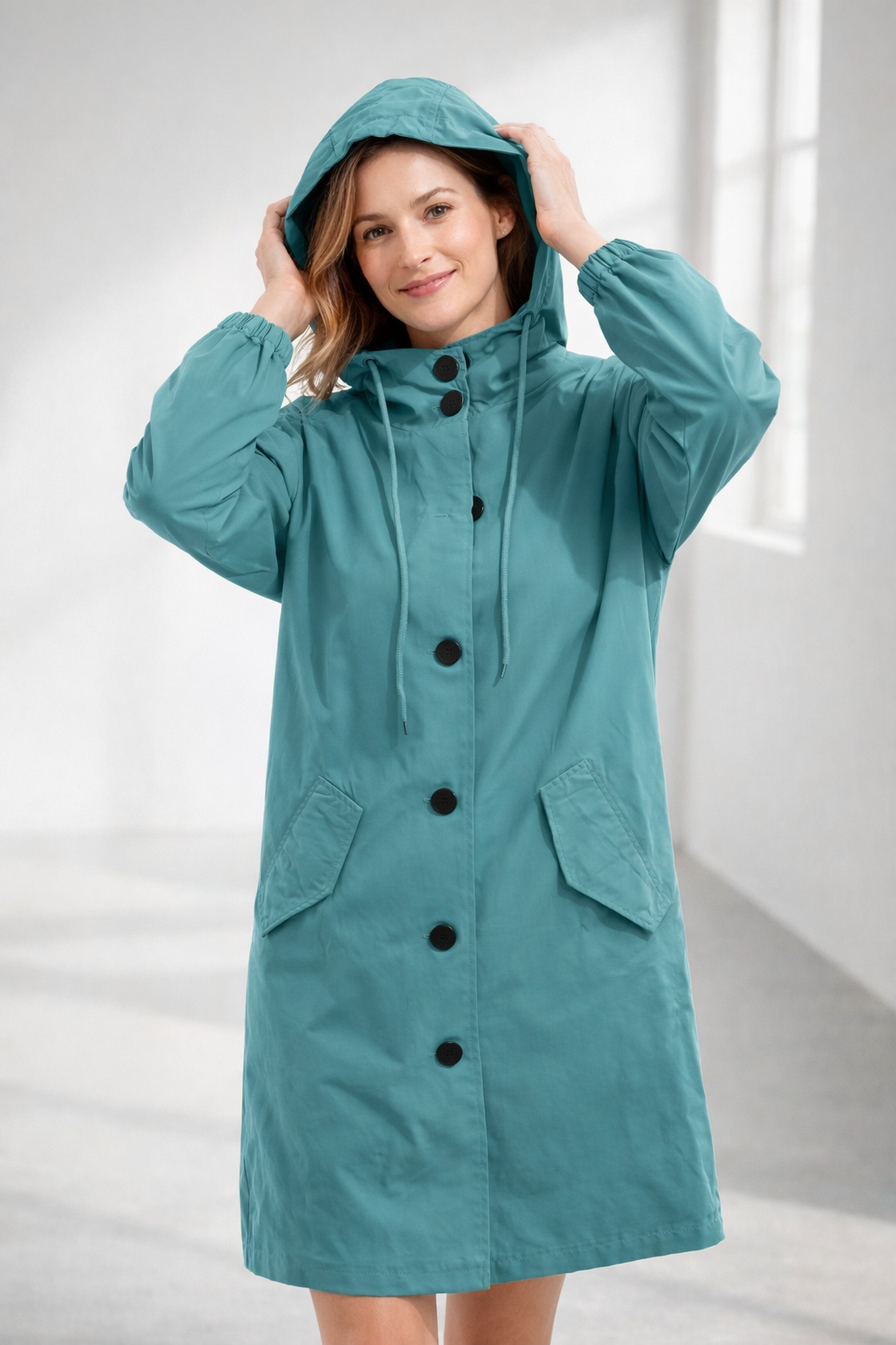 Pink - A woman wearing a teal mid-length hooded coat with a contrast black button-down front, angled flap pockets, and elasticated cuffs, set against a bright minimalist interior.