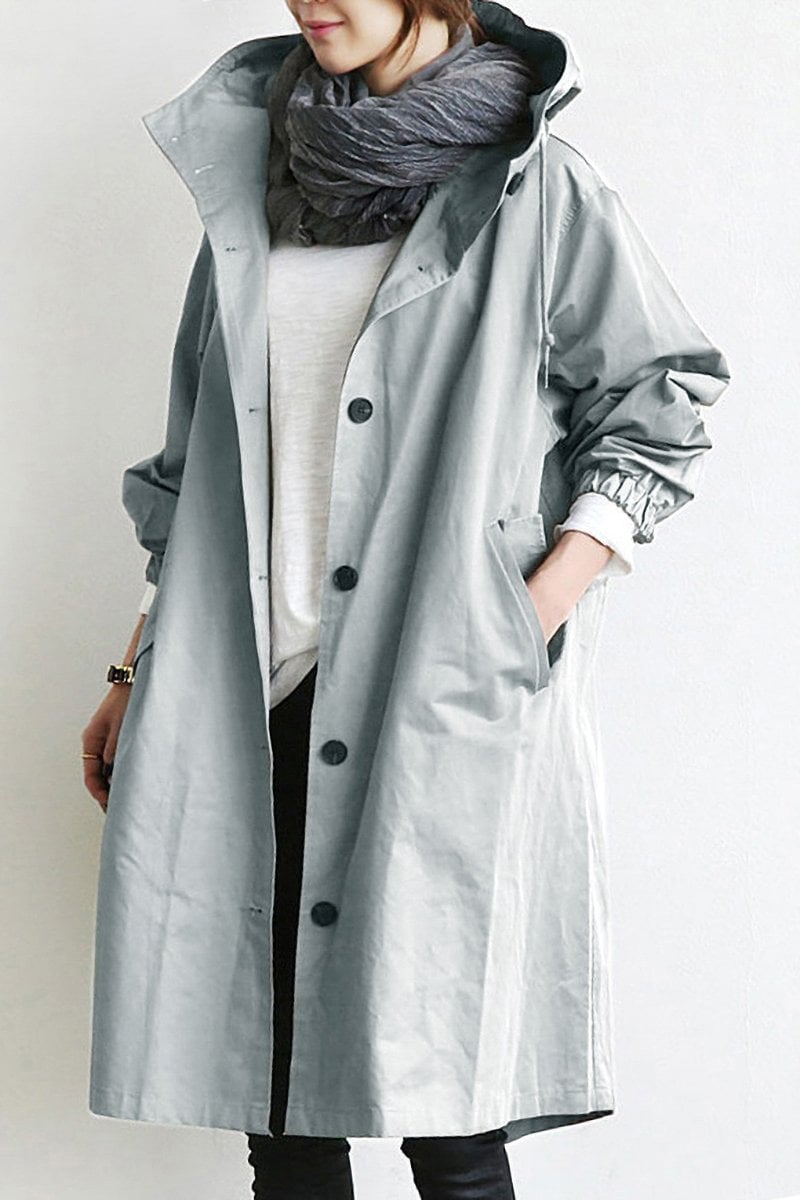 Pink - A woman wearing a teal mid-length hooded coat with a contrast black button-down front, angled flap pockets, and elasticated cuffs, set against a bright minimalist interior.