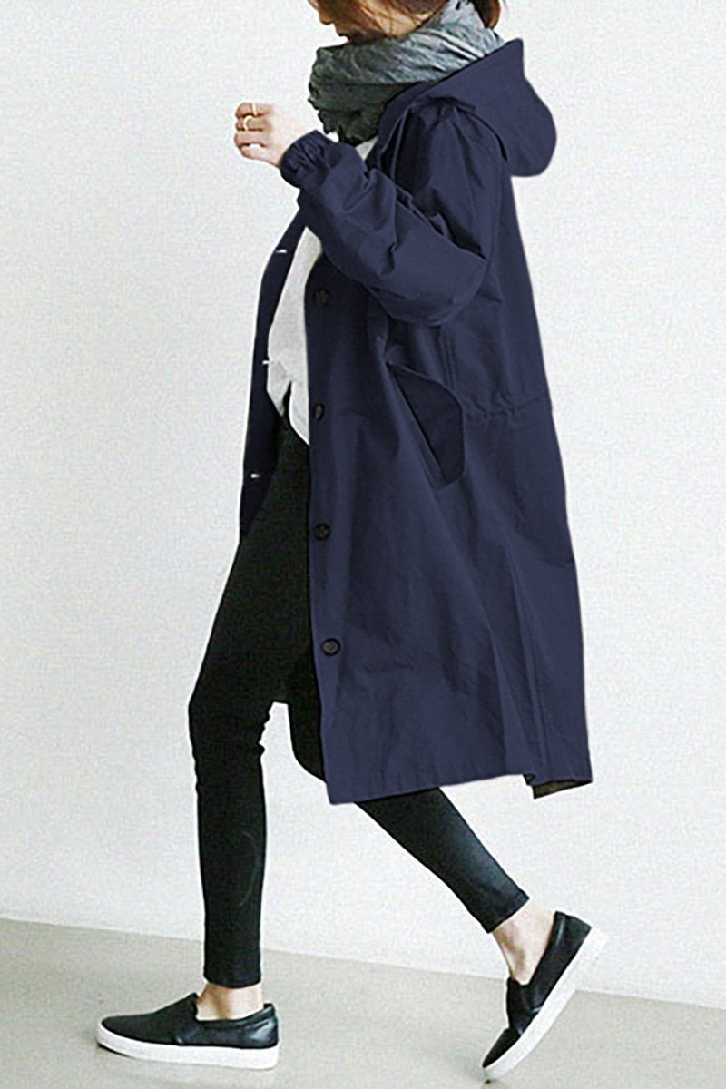 Pink - A woman wearing a teal mid-length hooded coat with a contrast black button-down front, angled flap pockets, and elasticated cuffs, set against a bright minimalist interior.