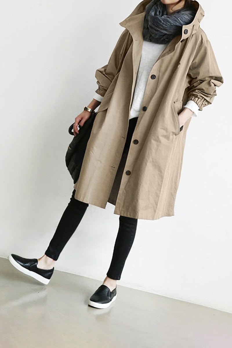 Pink - A woman wearing a teal mid-length hooded coat with a contrast black button-down front, angled flap pockets, and elasticated cuffs, set against a bright minimalist interior.