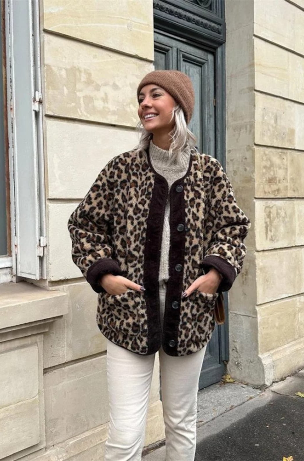 A woman wearing a relaxed-fit, fuzzy knit cardigan featuring a bold leopard print and contrasting dark ribbed placket, styled open over a white top and blue jeans.