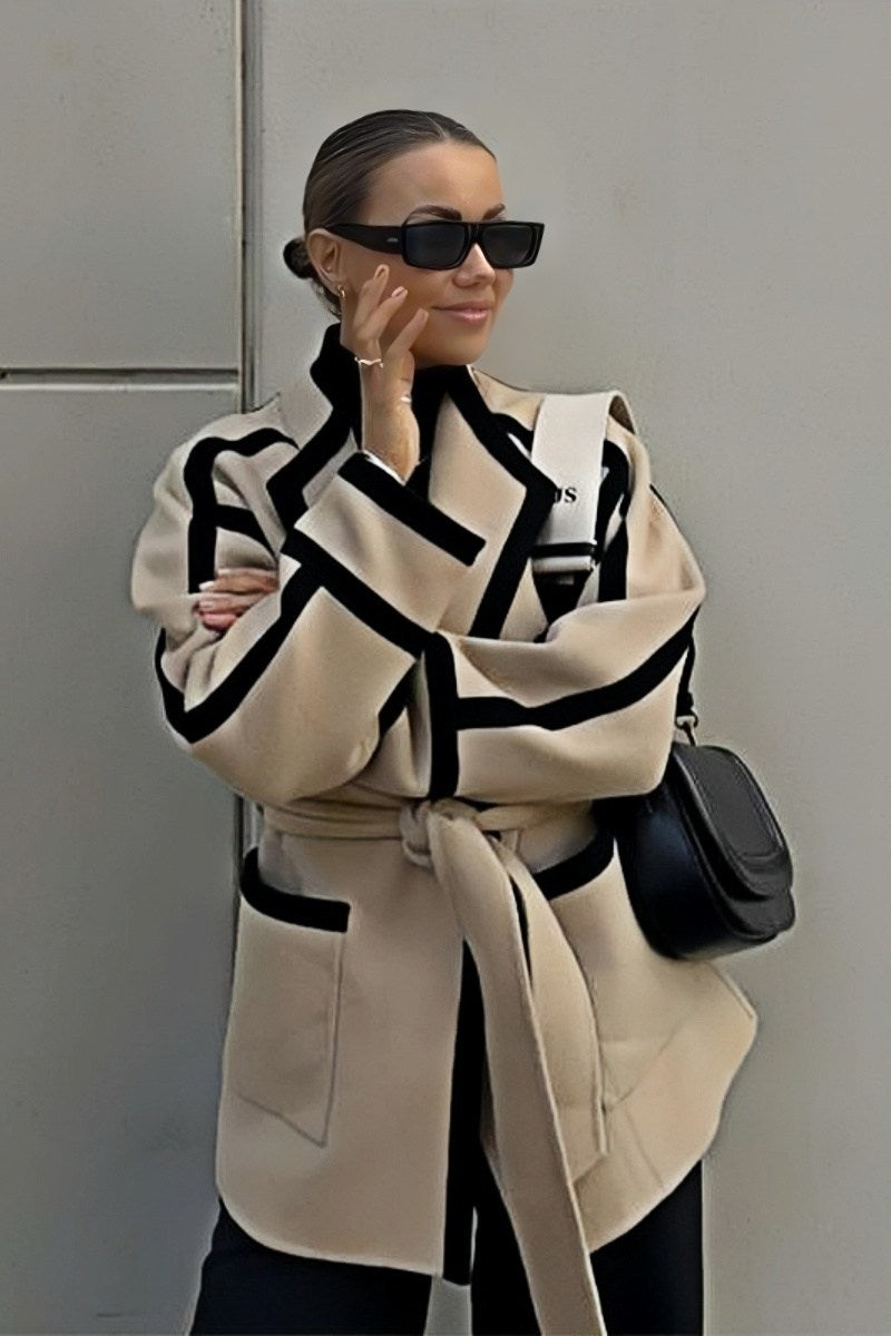 A woman wearing a tan collarless wrap jacket with bold black contrast trim and a self-tie belt, layered over a blue striped shirt and jeans.