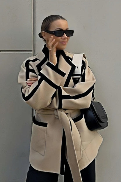 A woman wearing a tan collarless wrap jacket with bold black contrast trim and a self-tie belt, layered over a blue striped shirt and jeans.