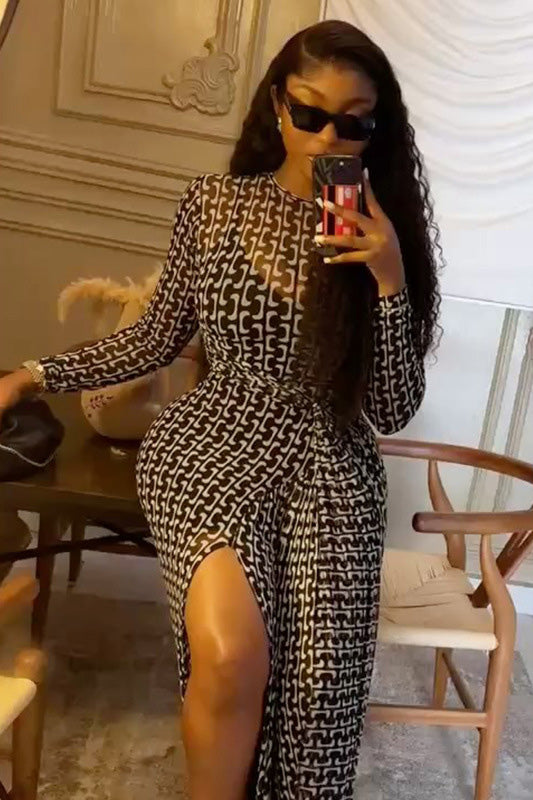 Black - A woman wearing a long-sleeved maxi dress featuring a black and white geometric chain link print, sheer mesh neckline, ruched waist detailing, and a high thigh slit.