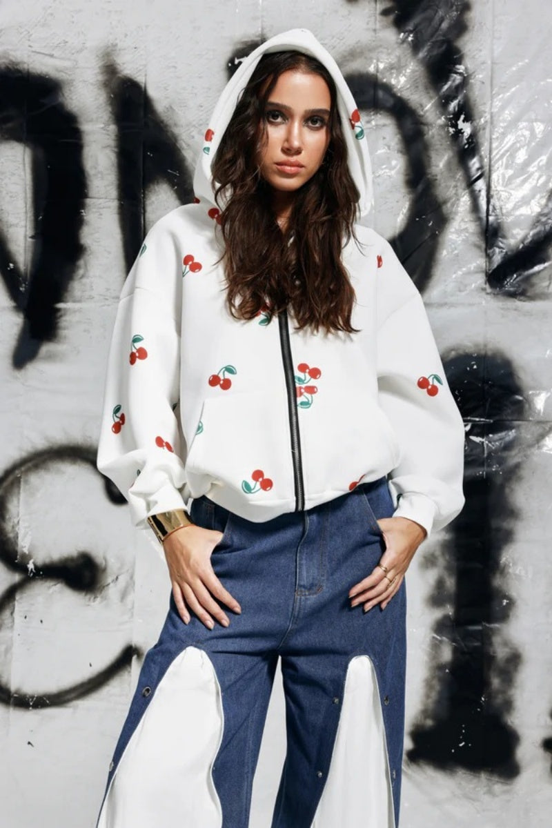 A woman wearing a white, oversized zip-up hoodie featuring a scattered red cherry print, paired with unique denim trousers.