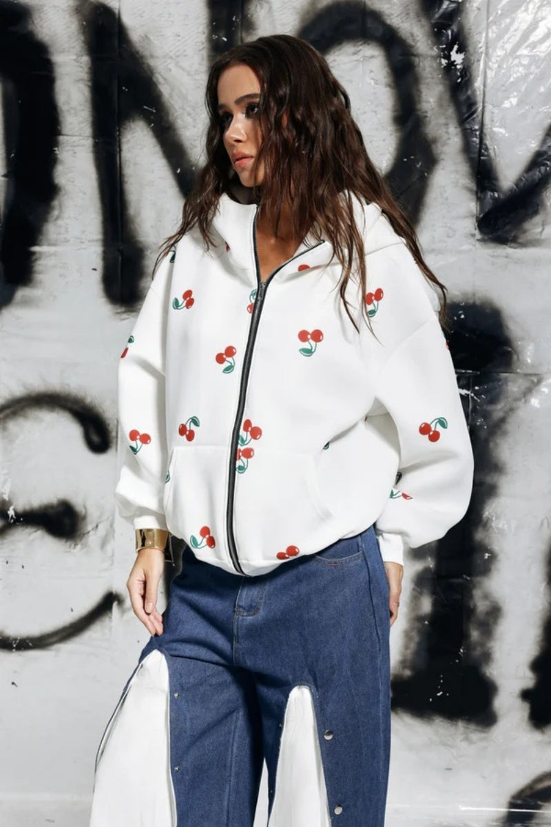 A woman wearing a white, oversized zip-up hoodie featuring a scattered red cherry print, paired with unique denim trousers.