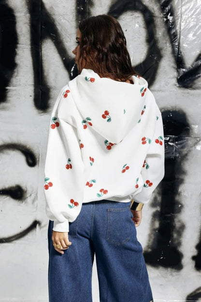White - A woman wearing a white, oversized zip-up hoodie featuring a scattered red cherry print, paired with unique denim trousers.