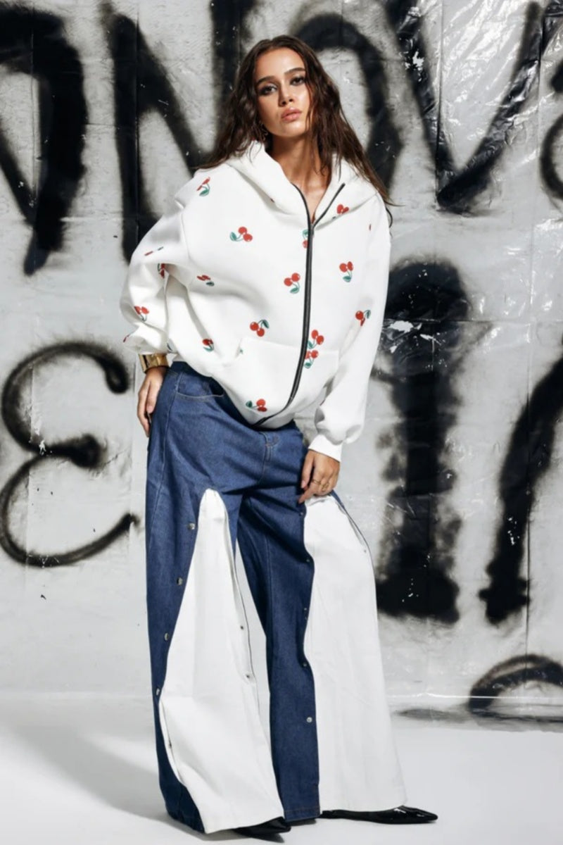 White - A woman wearing a white, oversized zip-up hoodie featuring a scattered red cherry print, paired with unique denim trousers.