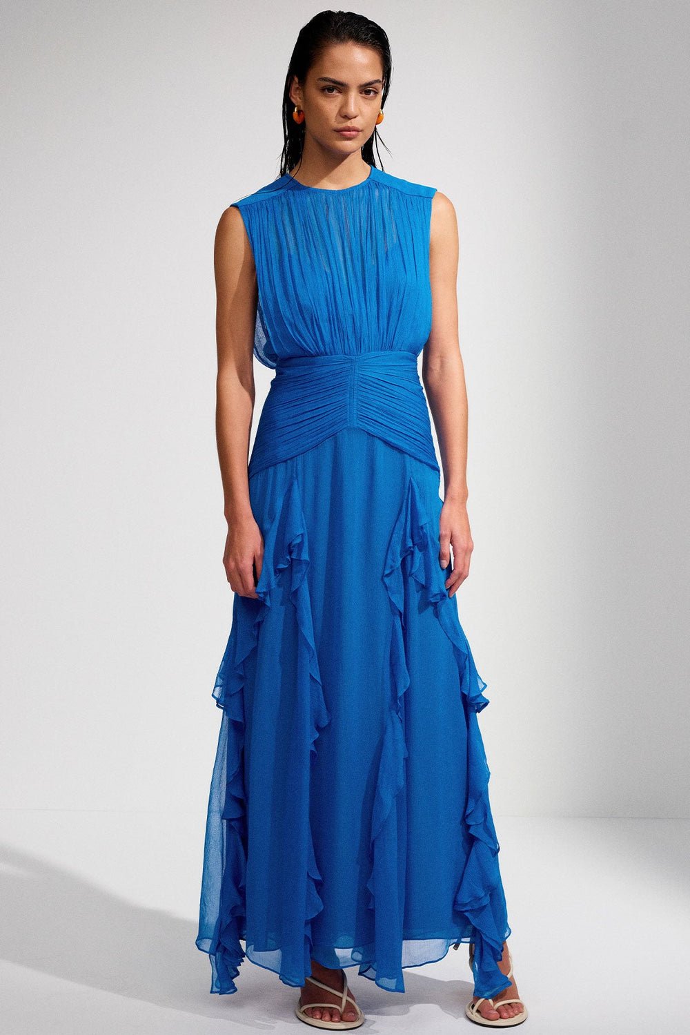 Blue - A woman wearing a vibrant cobalt blue sleeveless chiffon maxi dress featuring a high gathered neckline, a wide ruched waistband, and cascading vertical ruffles along the skirt.