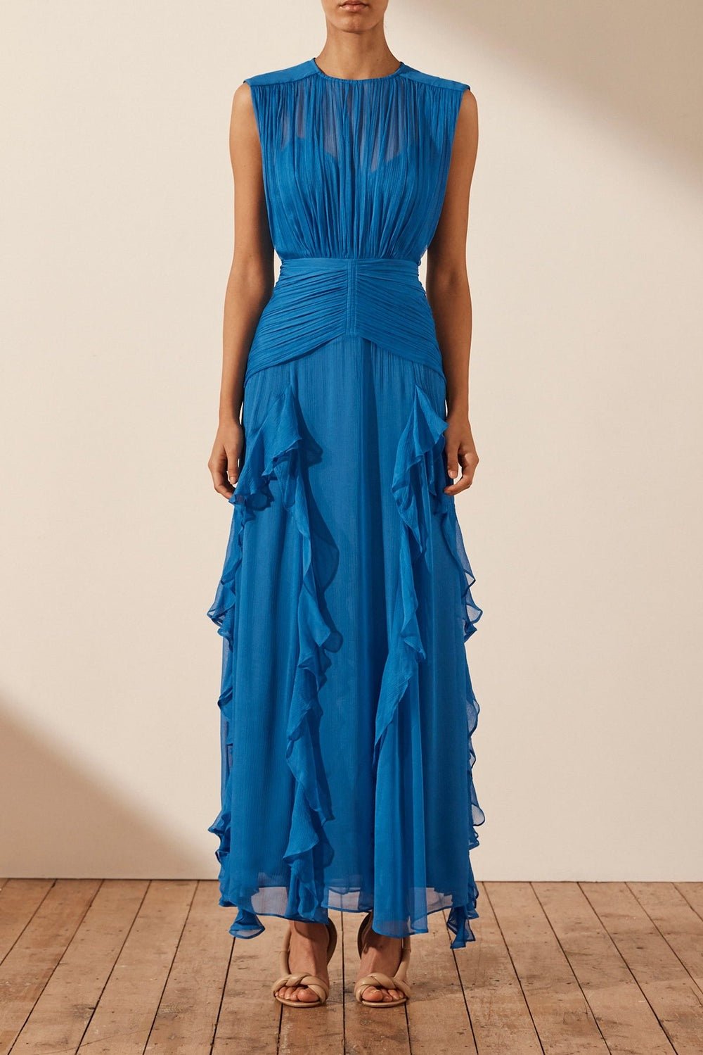 Blue - A woman wearing a vibrant cobalt blue sleeveless chiffon maxi dress featuring a high gathered neckline, a wide ruched waistband, and cascading vertical ruffles along the skirt.
