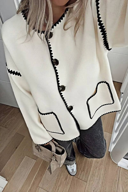 A woman wearing a cream-coloured knit cardigan featuring black contrast stitching along the edges, neckline, and front patch pockets, styled with dark denim jeans.