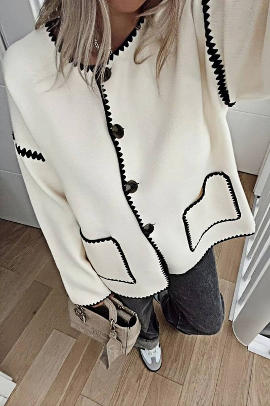 A woman wearing a cream-coloured knit cardigan featuring black contrast stitching along the edges, neckline, and front patch pockets, styled with dark denim jeans.