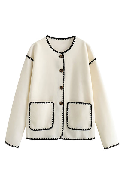 A woman wearing a cream-coloured knit cardigan featuring black contrast stitching along the edges, neckline, and front patch pockets, styled with dark denim jeans.