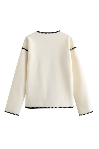 A woman wearing a cream-coloured knit cardigan featuring black contrast stitching along the edges, neckline, and front patch pockets, styled with dark denim jeans.
