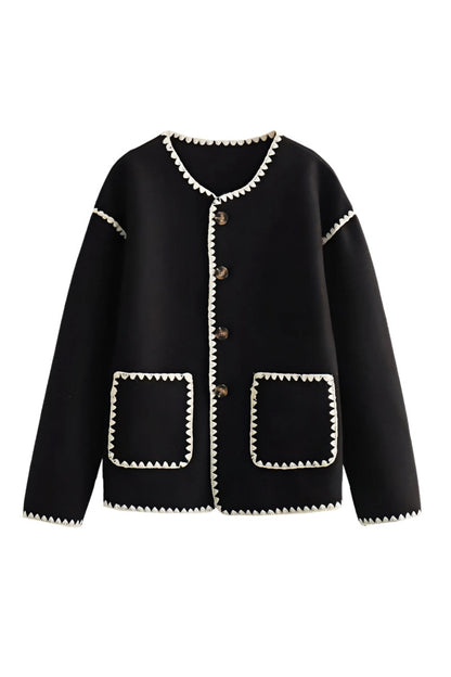 A woman wearing a cream-coloured knit cardigan featuring black contrast stitching along the edges, neckline, and front patch pockets, styled with dark denim jeans.