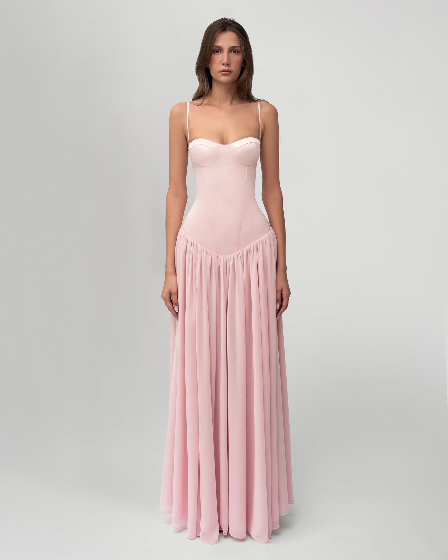 A woman wearing a light pink formal maxi dress featuring a structured corset bodice with spaghetti straps and a flowing, gathered skirt.