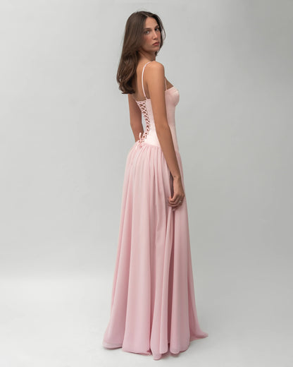 A woman wearing a light pink formal maxi dress featuring a structured corset bodice with spaghetti straps and a flowing, gathered skirt.