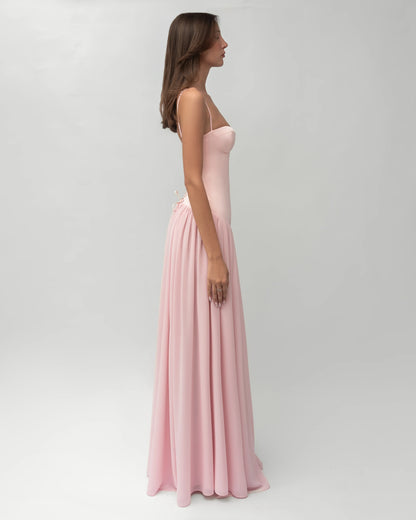 A woman wearing a light pink formal maxi dress featuring a structured corset bodice with spaghetti straps and a flowing, gathered skirt.