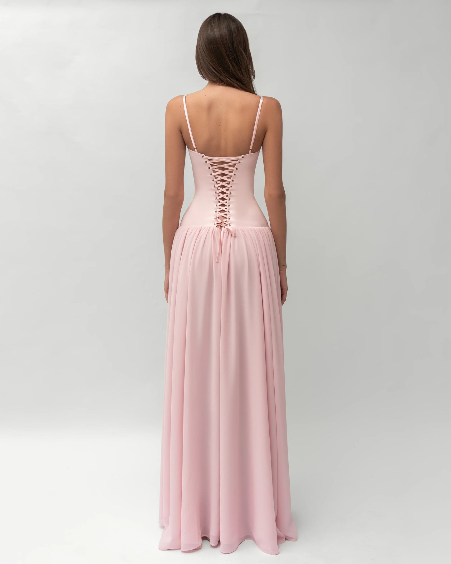 A woman wearing a light pink formal maxi dress featuring a structured corset bodice with spaghetti straps and a flowing, gathered skirt.