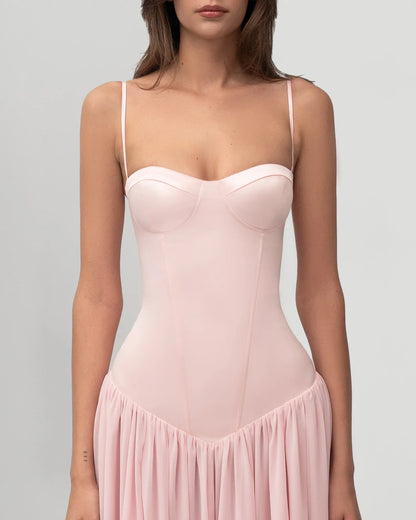 A woman wearing a light pink formal maxi dress featuring a structured corset bodice with spaghetti straps and a flowing, gathered skirt.