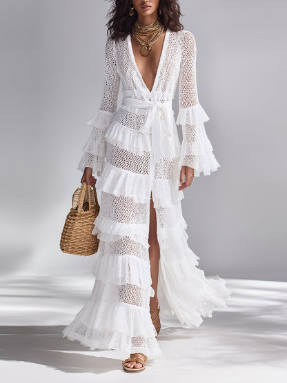 White - A woman wears a white crochet knit maxi dress with a deep V-neckline, long sleeves with tiered ruffles, and a tiered ruffled skirt. She holds a woven straw handbag.