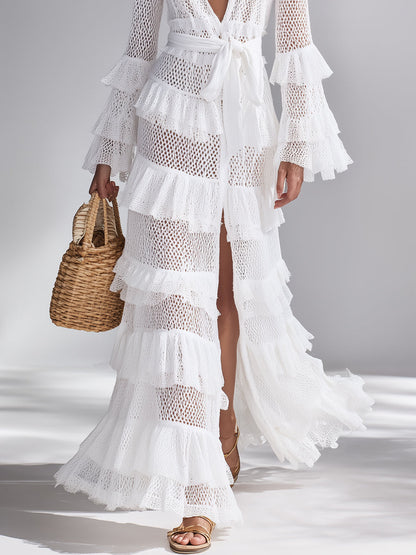 White - A woman wears a white crochet knit maxi dress with a deep V-neckline, long sleeves with tiered ruffles, and a tiered ruffled skirt. She holds a woven straw handbag.