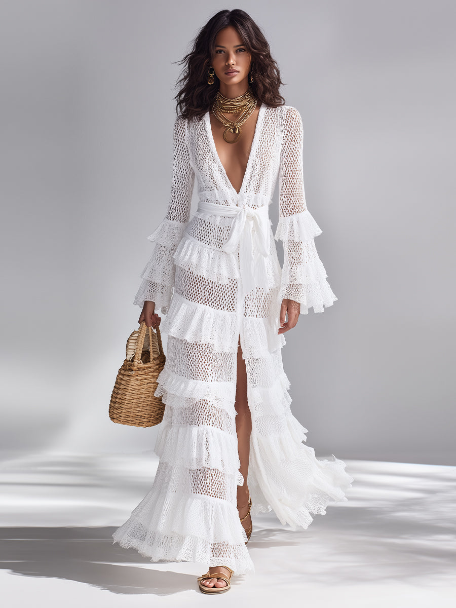White - A woman wears a white crochet knit maxi dress with a deep V-neckline, long sleeves with tiered ruffles, and a tiered ruffled skirt. She holds a woven straw handbag.