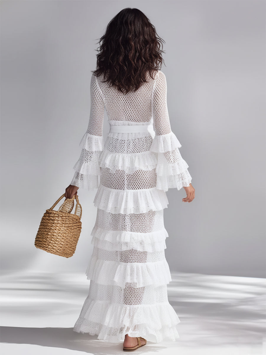 White - A woman wears a white crochet knit maxi dress with a deep V-neckline, long sleeves with tiered ruffles, and a tiered ruffled skirt. She holds a woven straw handbag.