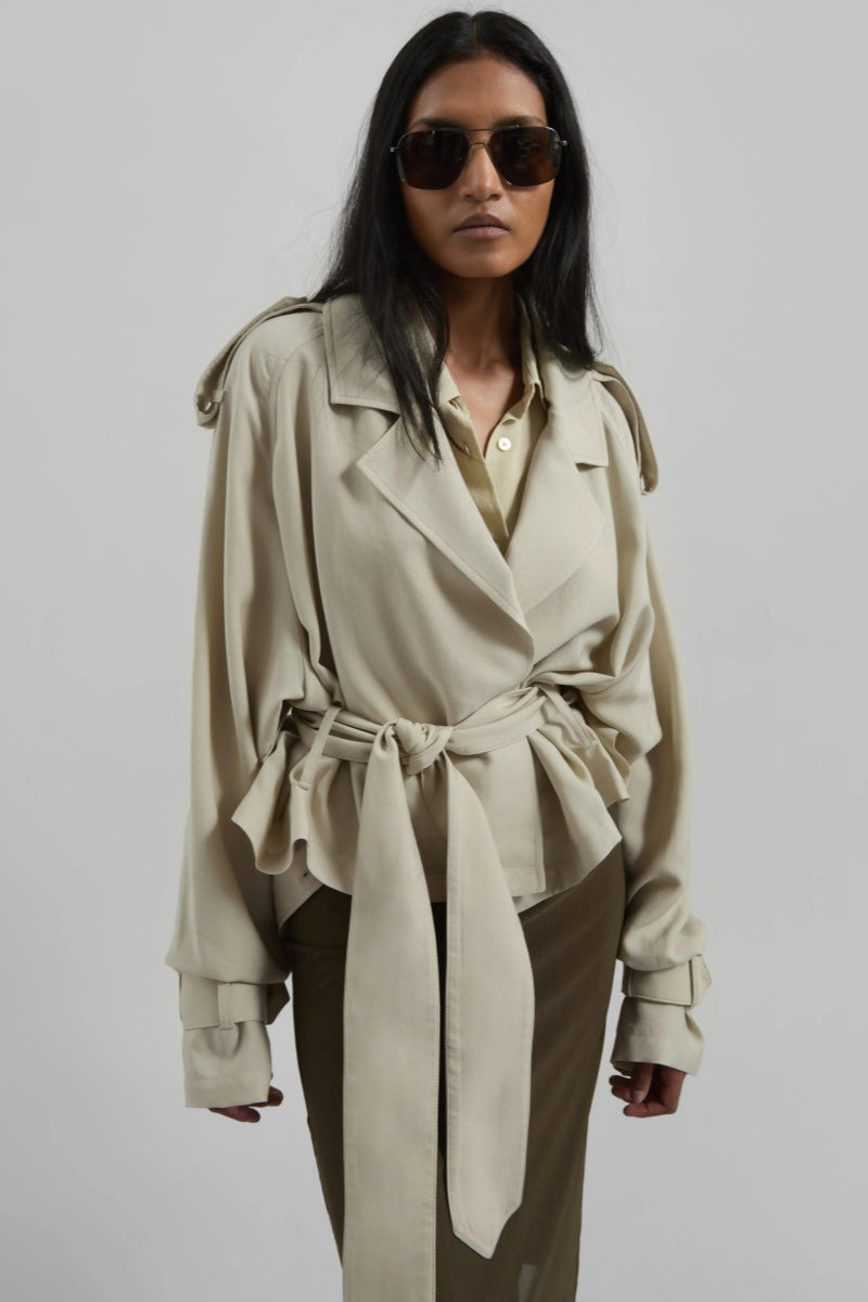 A model wearing a light beige cropped trench jacket with a belted waist, wide lapels, shoulder epaulettes, and relaxed sleeves.