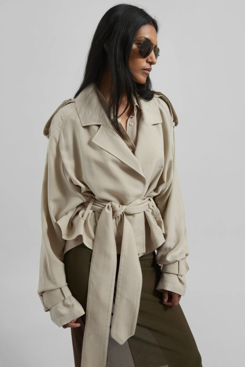 A model wearing a light beige cropped trench jacket with a belted waist, wide lapels, shoulder epaulettes, and relaxed sleeves.