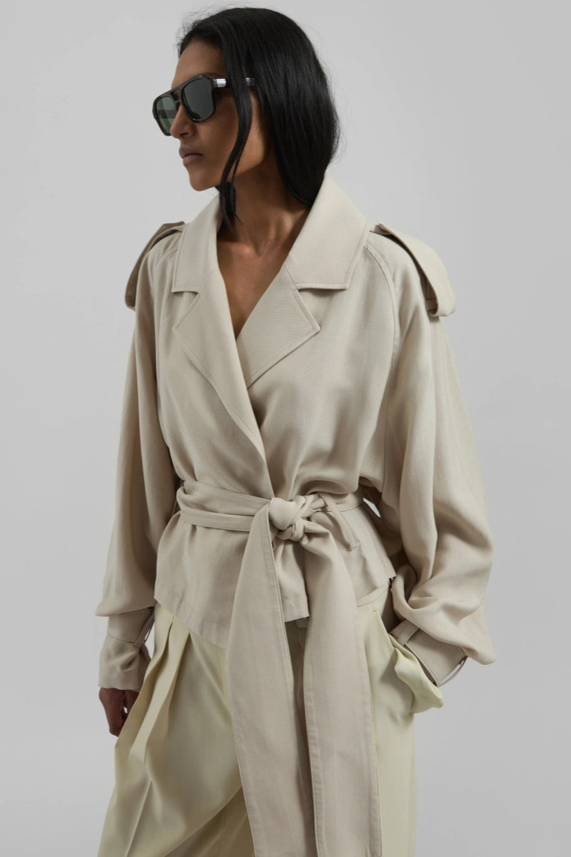 A model wearing a light beige cropped trench jacket with a belted waist, wide lapels, shoulder epaulettes, and relaxed sleeves.