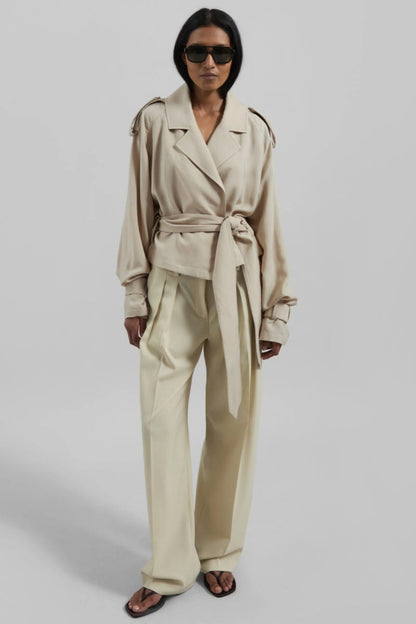A model wearing a light beige cropped trench jacket with a belted waist, wide lapels, shoulder epaulettes, and relaxed sleeves.