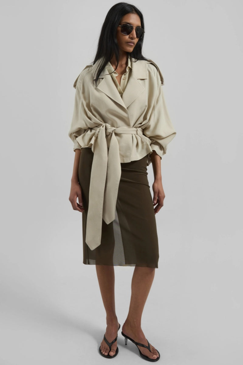 A model wearing a light beige cropped trench jacket with a belted waist, wide lapels, shoulder epaulettes, and relaxed sleeves.