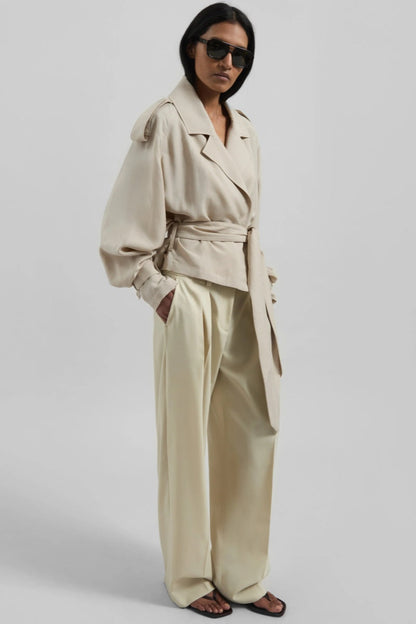 A model wearing a light beige cropped trench jacket with a belted waist, wide lapels, shoulder epaulettes, and relaxed sleeves.