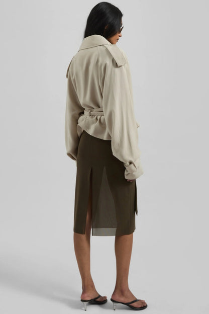 A model wearing a light beige cropped trench jacket with a belted waist, wide lapels, shoulder epaulettes, and relaxed sleeves.