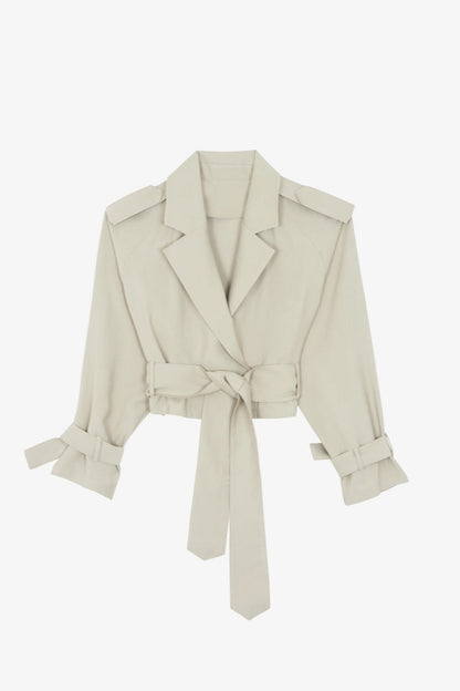 Cement - A model wearing a light beige cropped trench jacket with a belted waist, wide lapels, shoulder epaulettes, and relaxed sleeves.