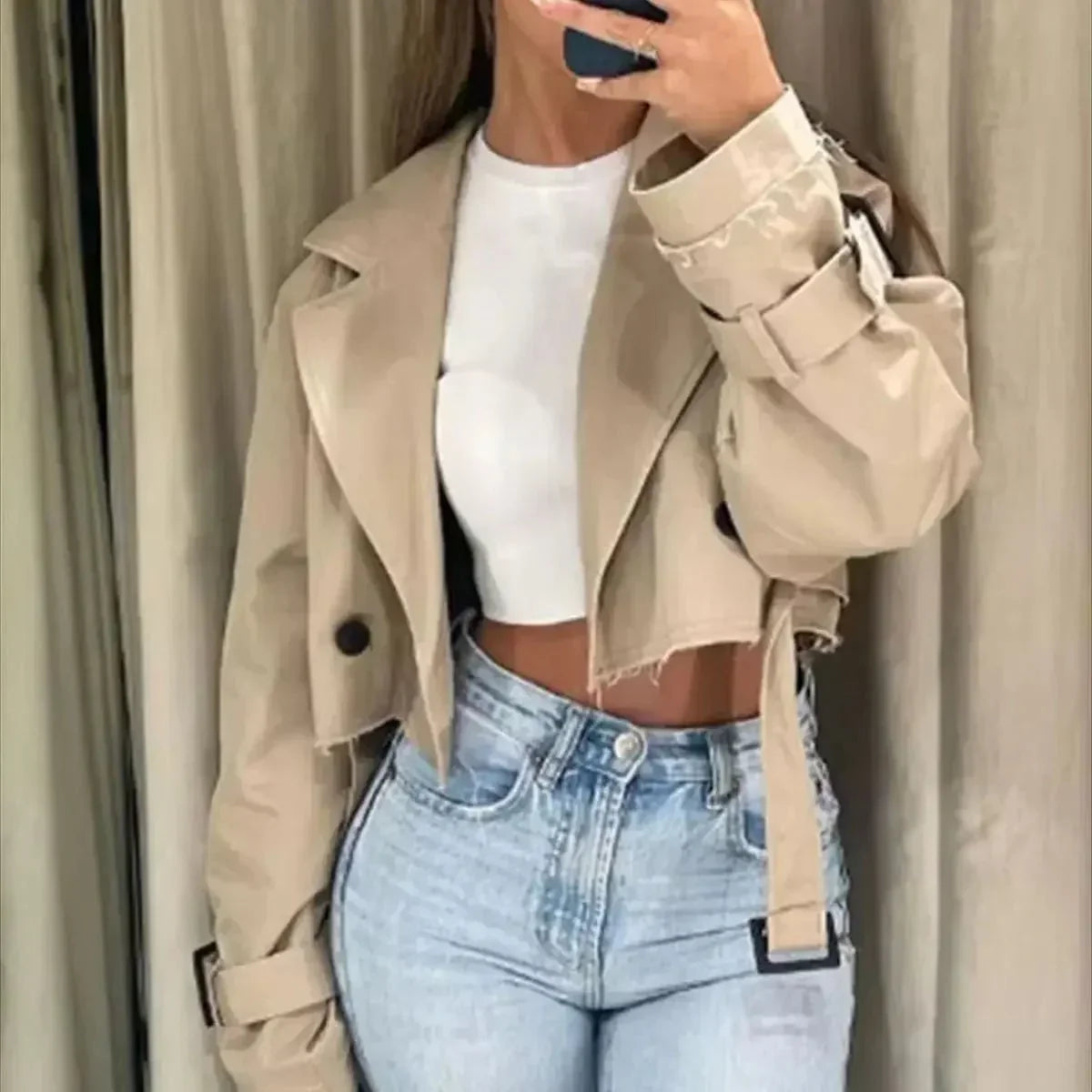 Women's beige cropped trench jacket featuring a frayed raw hem, wide lapels, and belted cuffs, styled open over a white top and blue jeans.