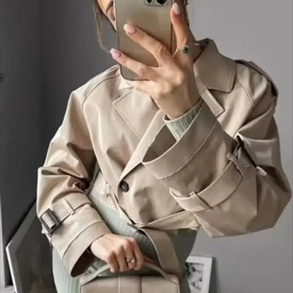 Women's beige cropped trench jacket featuring a frayed raw hem, wide lapels, and belted cuffs, styled open over a white top and blue jeans.