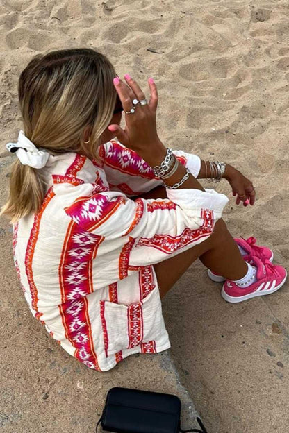 A woman wearing a relaxed, open-front kimono jacket featuring a vibrant pink and orange geometric Aztec print over a cream base, styled over a crochet crop top and shorts.